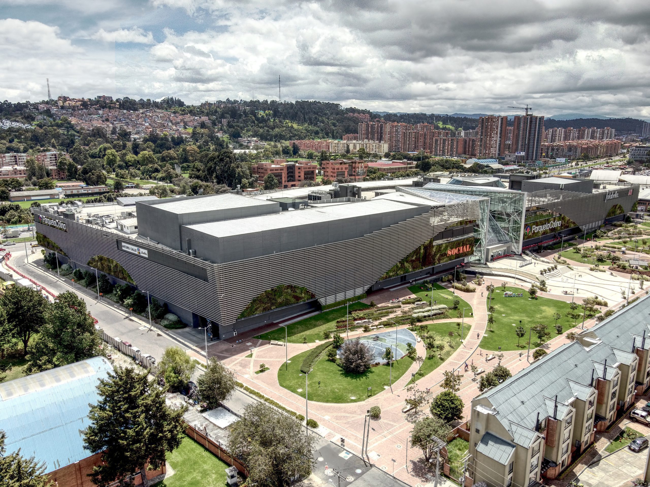 Fotografía aérea de un centro comercial en Bogotá, tomada desde un drone no tripulado. La imagen muestra una vista panorámica del edificio y su entorno, permitiendo ver los detalles de su arquitectura y su ubicación en la ciudad. Esta fotografía es una herramienta valiosa para promocionar el centro comercial y atraer a potenciales visitantes, así como para dar una idea general del área a aquellos que estén buscando servicios y tiendas en esa zona de la ciudad.